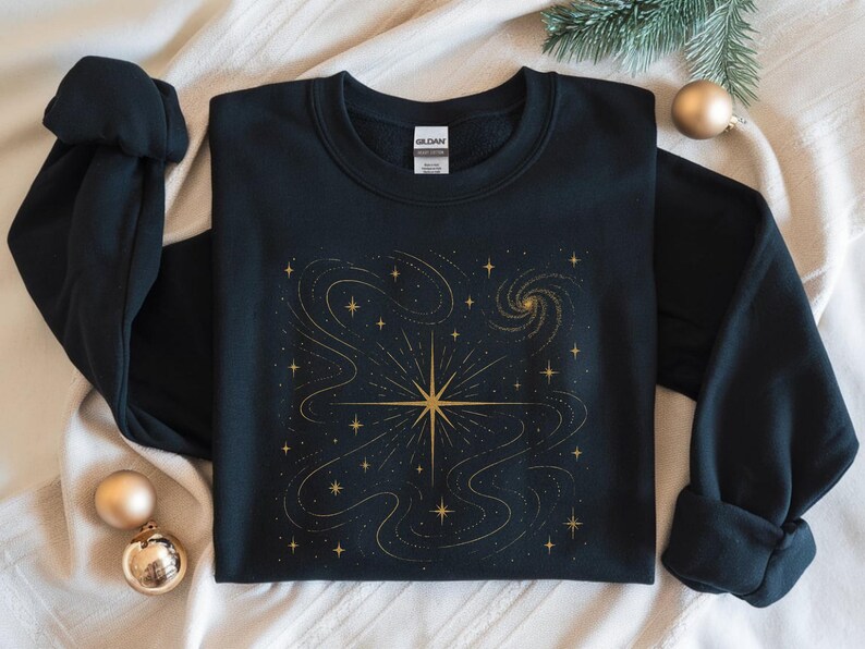 May include: Black sweatshirt featuring a gold celestial design. The graphic includes a central starburst, swirling lines, and numerous smaller stars. The design is centered on the front of the long-sleeved garment, creating a cosmic theme.