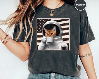 Könnte beinhalten: Dunkelgraues T-Shirt mit einem Aufdruck einer orangefarbenen Katze in einem Raumanzug, die eine Tasse hält, vor dem Hintergrund einer amerikanischen Flagge. Die Katze hat einen ernsten Gesichtsausdruck. Das T-Shirt ist ein bequemer, lässiger Stil.