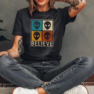 Op de afbeelding: Zwart T-shirt met een retro design. Het heeft vier alien gezichten in turquoise, witte, gele en oranje vierkanten. Het woord "BELIEVE" staat onder de alien gezichten in een vintage lettertype.