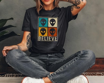 Puede incluir: Camiseta negra con un diseño retro. Presenta cuatro caras de extraterrestres en cuadrados turquesa, blancos, amarillos y naranjas. La palabra "BELIEVE" está impresa debajo de las caras de los extraterrestres en una fuente vintage.