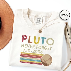 Könnte beinhalten: Elfenbeinfarbenes T-Shirt mit dem Text "PLUTO NEVER FORGET 1930-2006" in bunten Buchstaben. Das Design beinhaltet eine Grafik von Pluto und eine Reihe horizontaler Streifen in verschiedenen Farben. Eine goldene Halskette liegt über dem Shirt.