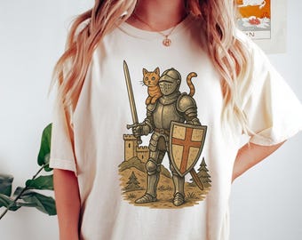 May include: Cream-colored t-shirt featuring a cartoon knight in silver armor, holding a sword and shield with a cross. An orange tabby cat sits on the knight's shoulder. A castle and trees are in the background.
