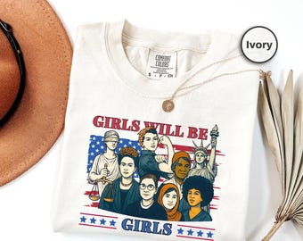 May include: Ivory-colored t-shirt featuring a graphic of diverse women with the text "GIRLS WILL BE GIRLS" in red and blue. The design includes illustrations of historical figures and the American flag.