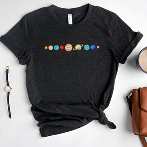 May include: A dark gray t-shirt with a graphic of the planets in our solar system in a line across the chest. The planets are depicted in their natural colors.