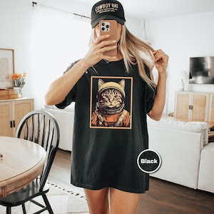 May include: Black t-shirt featuring a cat in an astronaut helmet and suit, framed in a gold border. The shirt is worn with a black baseball cap that says "COWBOY HAT". The shirt is a comfortable, casual style.