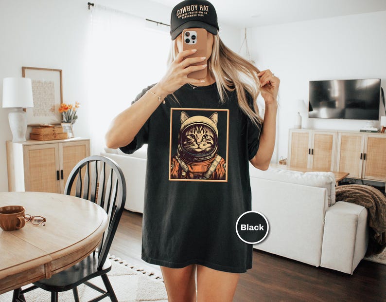May include: Black t-shirt featuring a cat in an astronaut helmet and suit, framed in a gold border. The shirt is worn with a black baseball cap that says "COWBOY HAT". The shirt is a comfortable, casual style.