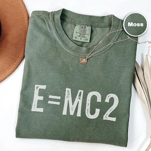 May include: A moss green t-shirt with the equation E=MC2 printed in white. The shirt has a crew neck and a small tag on the collar. A gold necklace with a pendant is draped across the shirt.