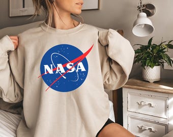 May include: Beige crewneck sweatshirt featuring the NASA logo. The logo is a blue circle with white text and a red swoosh. The sweatshirt is a neutral color, suitable for casual wear.