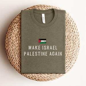 Op de afbeelding: Olijfgroen t-shirt met de witte tekst "MAKE ISRAEL PALESTINE AGAIN" in een verweerde lettertype. Het shirt heeft een kleine Palestijnse vlag boven de tekst. Gepresenteerd op een geweven, cirkelvormig oppervlak.