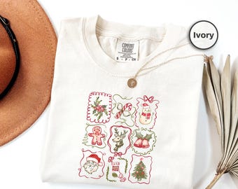 Puede incluir: Camiseta Comfort Colors de color marfil con una cuadrícula de ilustraciones navideñas. Las ilustraciones incluyen un muñeco de nieve, Papá Noel, un hombre de jengibre, un reno y un árbol de Navidad. Un collar dorado está drapeado sobre la camisa.
