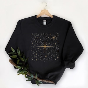 May include: Black sweatshirt with a gold star and celestial design. The graphic features a central starburst with swirling lines and smaller stars, creating a cosmic theme. The sweatshirt is hanging on a wooden hanger.