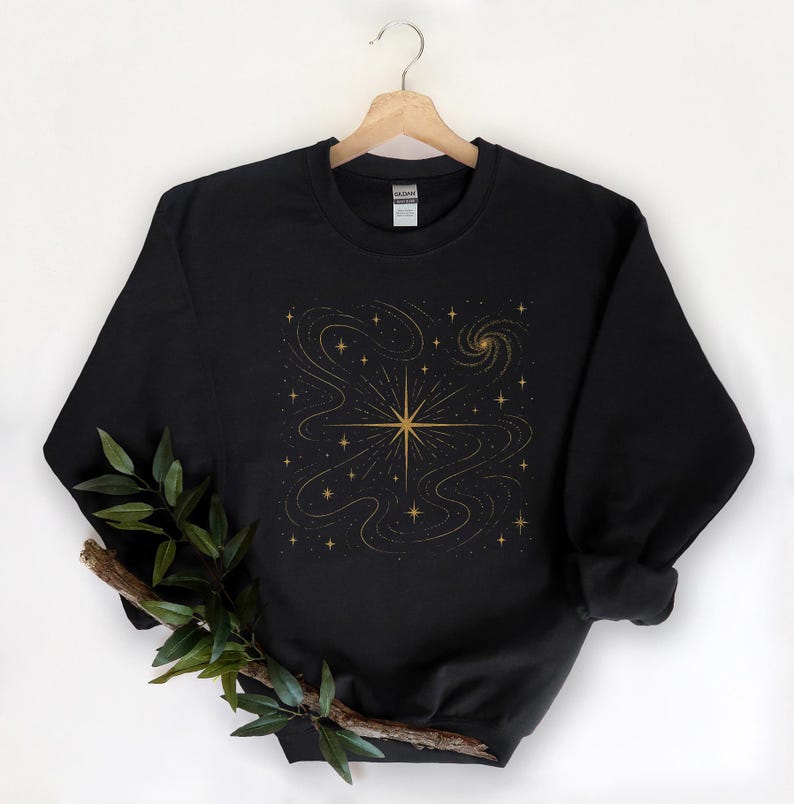 May include: Black sweatshirt with a gold star and celestial design. The graphic features a central starburst with swirling lines and smaller stars, creating a cosmic theme. The sweatshirt is hanging on a wooden hanger.