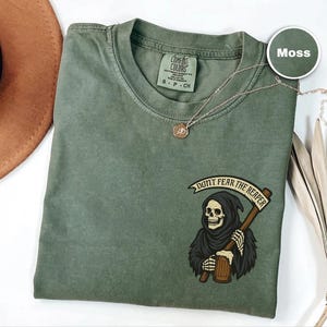 Puede incluir: Camiseta verde musgo con un gr&aacute;fico de la Parca sosteniendo una guada&ntilde;a y el texto "DON'T FEAR THE REAPER". La camiseta tiene cuello redondo y mangas cortas. Tambi&eacute;n se ve un collar dorado y un sombrero marr&oacute;n.