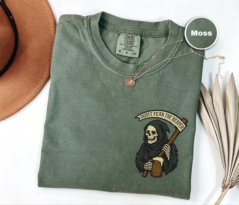 Puede incluir: Camiseta verde musgo con un gr&aacute;fico de la Parca sosteniendo una guada&ntilde;a y el texto "DON'T FEAR THE REAPER". La camiseta tiene cuello redondo y mangas cortas. Tambi&eacute;n se ve un collar dorado y un sombrero marr&oacute;n.