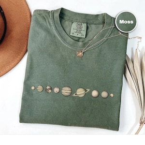 May include: A moss green t-shirt featuring a line of planets across the front. The planets are in shades of brown and beige. A silver necklace with a small pendant is on the shirt. The shirt is folded on a white surface.