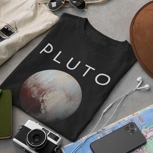May include: Black t-shirt with a white graphic of Pluto, the dwarf planet, and the word "PLUTO" in white text.