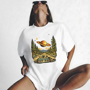 May include: White t-shirt with a graphic of a car on a forest road, with a planet in the sky. The design includes trees, stars, and mountains. The shirt is a casual, unisex style, suitable for everyday wear.