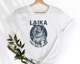 Könnte beinhalten: Weißes T-Shirt mit einer marineblauen Grafik eines Hundes in einem Raumanzug, mit dem Text "LAIKA FIRST DOG IN SPACE". Die Hundeillustration befindet sich in einem marineblauen Kreis mit Sternen. Das T-Shirt hängt an einem Holzbügel.