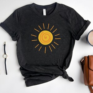 May include: Dark gray t-shirt featuring a yellow sun graphic. The sun design has a spiral center and radiating lines. The shirt is styled with rolled-up sleeves and a knotted hem. Accessories include a watch, glasses, and a brown purse.