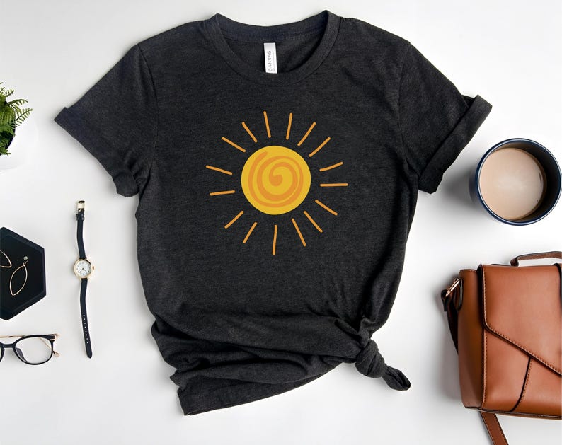 May include: Dark gray t-shirt featuring a yellow sun graphic. The sun design has a spiral center and radiating lines. The shirt is styled with rolled-up sleeves and a knotted hem. Accessories include a watch, glasses, and a brown purse.