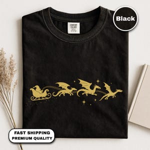 Pode incluir: Camiseta preta com uma ilustração dourada do Papai Noel em um trenó puxado por dragões. A camiseta tem gola redonda e uma etiqueta "Comfort Colors". As palavras "Fast Shipping Premium Quality" estão na parte inferior. A palavra "Black" está em um círculo.