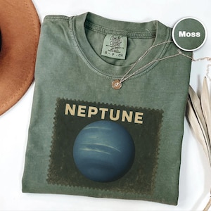 May include: A moss green t-shirt with a graphic of the planet Neptune. The word "NEPTUNE" is printed above a blue planet image. A gold necklace with a pendant is draped across the shirt. The shirt is a Comfort Colors brand.