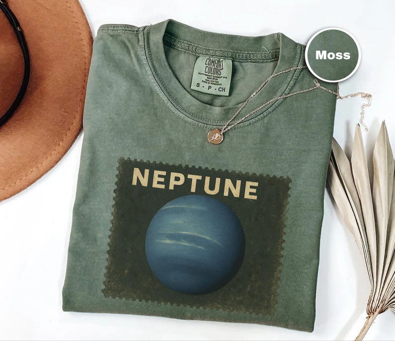 May include: A moss green t-shirt with a graphic of the planet Neptune. The word "NEPTUNE" is printed above a blue planet image. A gold necklace with a pendant is draped across the shirt. The shirt is a Comfort Colors brand.
