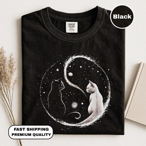 Könnte beinhalten: Schwarzes T-Shirt mit Yin-und-Yang-Design, das zwei Katzen zeigt, eine schwarze und eine weiße, vor einem Sternenhintergrund. Das T-Shirt hat einen Rundhalsausschnitt und ist aus weichem Stoff.