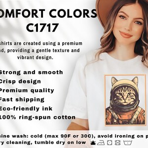 May include: White t-shirt with a framed illustration of a cat in an astronaut helmet. The text "COMFORT COLORS C1717" is at the top. The shirt is described as strong, smooth, and made with eco-friendly ink and 100% ring-spun cotton.