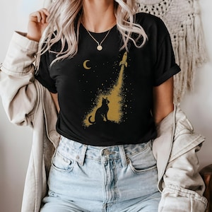 Puede incluir: Camiseta negra con un diseño dorado de un gato mirando un cohete que despega hacia el espacio, con una luna creciente y estrellas. El diseño está impreso en una camiseta negra.