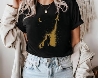 May include: Black t-shirt featuring a gold graphic of a cat gazing at a rocket launching into space, with a crescent moon and stars. The design is printed on a black t-shirt.