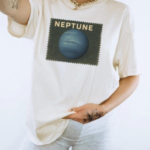 May include: Cream-colored t-shirt featuring a graphic of the planet Neptune within a postage stamp design. The word "NEPTUNE" is printed above the planet. The shirt is worn with gray shorts.