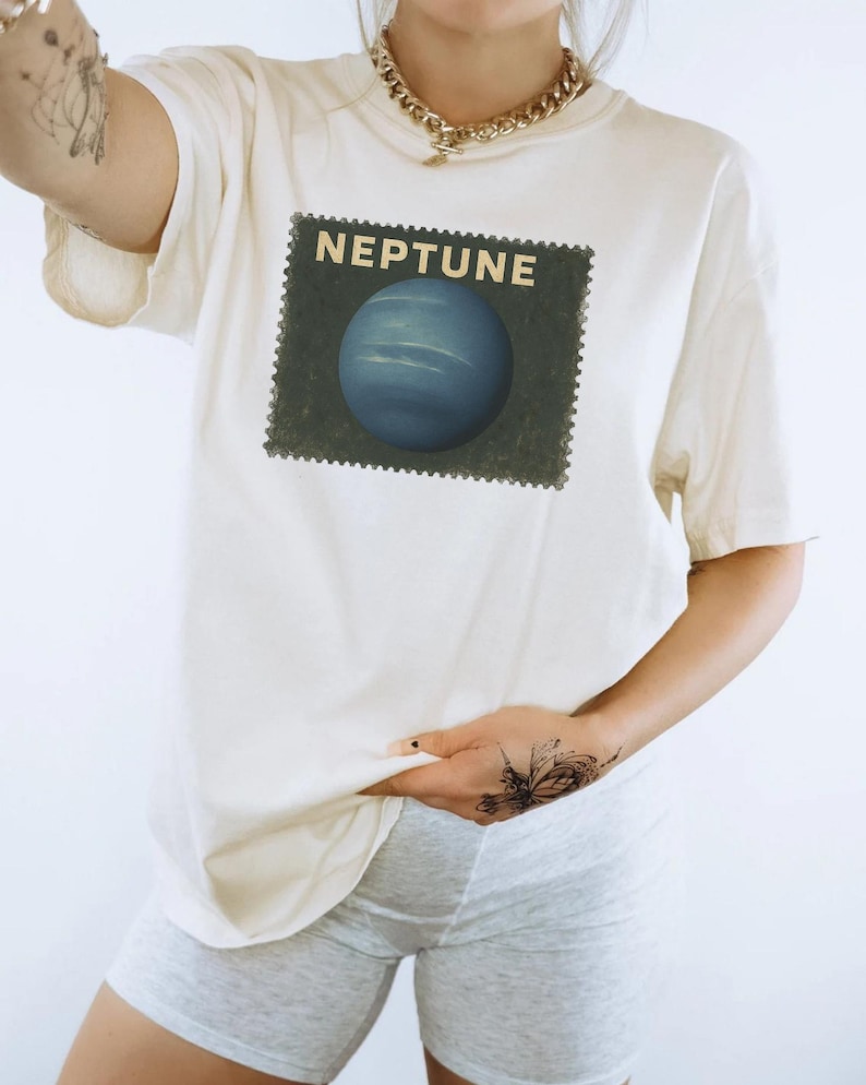 May include: Cream-colored t-shirt featuring a graphic of the planet Neptune within a postage stamp design. The word "NEPTUNE" is printed above the planet. The shirt is worn with gray shorts.