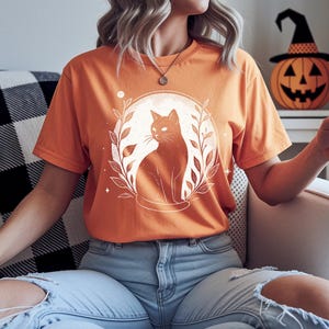 May include: Orange t-shirt featuring a black cat illustration within a circular design of leaves and a moon. The cat is sitting, facing right. The shirt is short-sleeved and has a crew neck. A Halloween-themed pumpkin with a witch hat is in the background.