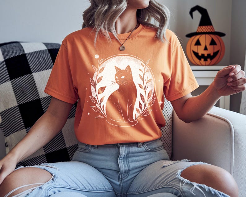 May include: Orange t-shirt featuring a black cat illustration within a circular design of leaves and a moon. The cat is sitting, facing right. The shirt is short-sleeved and has a crew neck. A Halloween-themed pumpkin with a witch hat is in the background.
