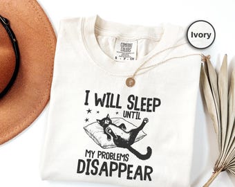 May include: Ivory-colored t-shirt with the text "I WILL SLEEP UNTIL MY PROBLEMS DISAPPEAR." A black cat is illustrated lying on a pillow. The shirt has a crew neck and short sleeves. A gold necklace is draped across the shirt.