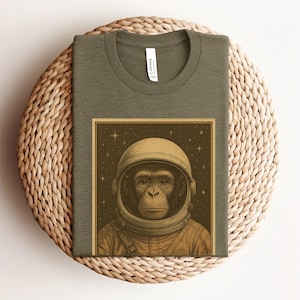 May include: A green t-shirt featuring a vintage-style illustration of a chimpanzee wearing an astronaut helmet. The illustration is surrounded by a brown frame and a starry background.