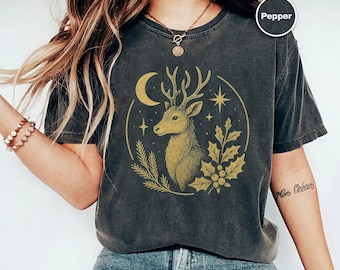 Puede incluir: Camiseta gris oscuro con un diseño de ciervo dorado dentro de un círculo, que incluye una luna creciente, estrellas y follaje. La palabra "Pepper" está en un círculo blanco. La camiseta es de estilo casual y cómodo.