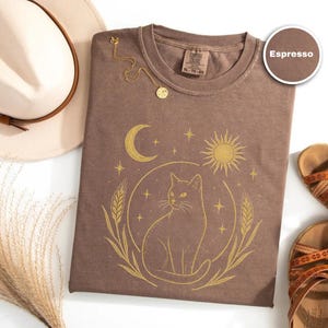 May include: Espresso-colored t-shirt with a gold cat design, a crescent moon, and a sun. The shirt is paired with a cream-colored hat, a gold necklace, and brown sandals. The shirt is XL.