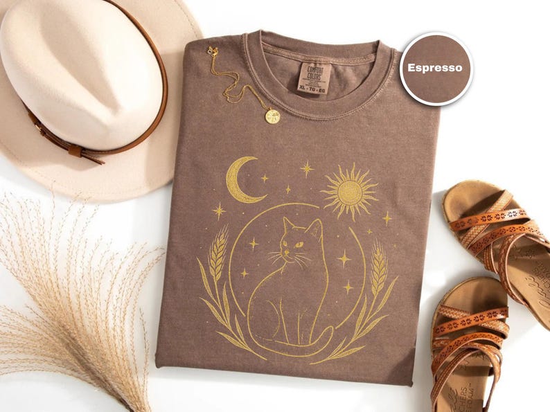 May include: Espresso-colored t-shirt with a gold cat design, a crescent moon, and a sun. The shirt is paired with a cream-colored hat, a gold necklace, and brown sandals. The shirt is XL.