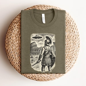 May include: Olive green t-shirt featuring a black and white illustration of a Roman soldier looking up at a UFO hovering over the Colosseum. The shirt has a crew neck and the brand name "Canvas" is visible.