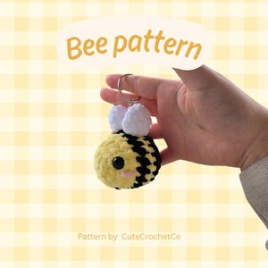Peut inclure: Un porte-clés en forme d'abeille crochetée, avec un corps jaune et noir, des ailes en pompons blancs et un anneau en métal. L'abeille a un visage souriant avec des yeux noirs et des joues roses. Le texte "Bee pattern" est dans une bulle jaune.