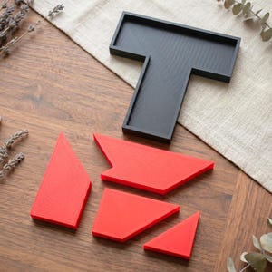 May include: A black, T-shaped tray with a textured surface sits on a wooden table. Four red geometric puzzle pieces are scattered around the tray. The pieces are various shapes and sizes, with a textured surface.