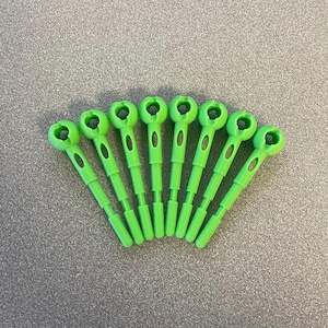 Puede incluir: Un abanico de siete protectores de puntas de agujas de tejer de plástico verde brillante. Cada protector tiene una parte superior redondeada con un agujero y un tallo largo y delgado con un recorte ovalado. Los protectores están dispuestos en semicírculo.