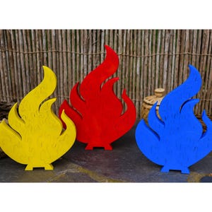 May include: Three wooden flame-shaped puzzles in yellow, red, and blue. Each puzzle is a different color and stands upright on a flat base. The background features a woven bamboo fence.