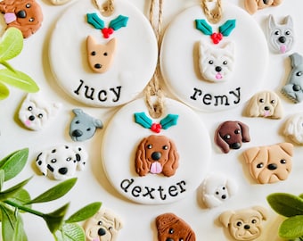 May include: White ceramic dog ornaments with names "Lucy", "Remy", and "Dexter" in black script. Each ornament features a dog face and holly accents. Additional dog face decorations surround the ornaments, creating a festive theme.
