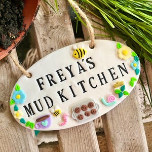 May include: A white, oval ceramic sign with the words "FREYA'S MUD KITCHEN" in black. The sign is adorned with colourful clay decorations like bees, flowers, a snail, and a tray of cookies. It is suspended by a natural twine.