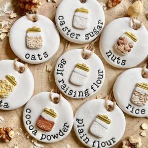 May include: Circular white ceramic pantry labels with jar illustrations and text labels including oats, pasta, caster sugar, nuts, self-raising flour, cocoa powder, plain flour, and rice. Each has a jute string.