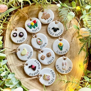 May include: A collection of 12 white ceramic Easter decorations with various designs and names. The decorations are decorated with images of a bunny, a chick, a lamb, a basket of eggs, a daffodil, a hot cross bun, and a chocolate egg. The decorations are strung with twine and are arranged on a wooden surface.