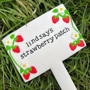 May include: White garden marker with the text "Lindsay's strawberry patch" and red and green strawberry and white flower decorations.
